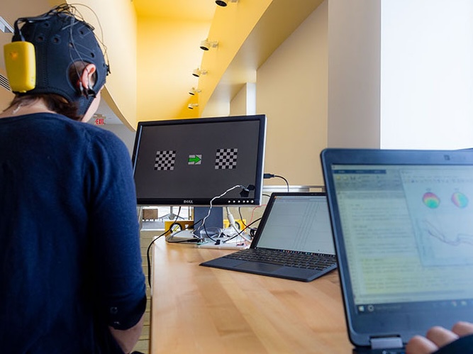 A person wearing an EEG cap sits at a desk facing a monitor displaying checkerboard patterns, while another person uses a laptop displaying brainwave data.