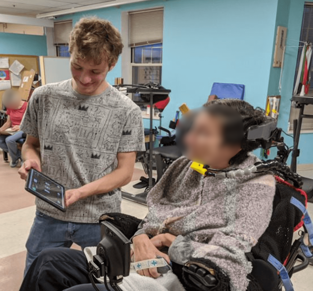 A man holds a tablet with a graphical design in front of a blurred-face powered wheelchair user using a sip-and-puff control system.