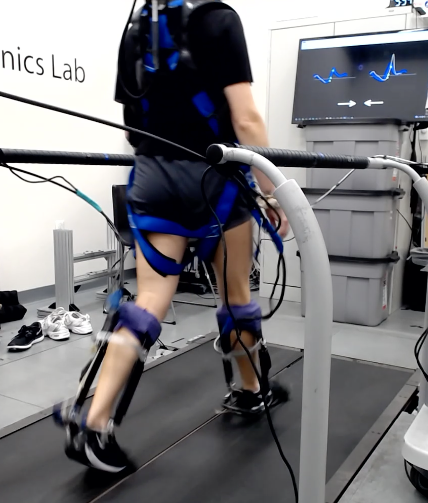 A person walks on a treadmill with ankle exoskeletons in a laboratory setting. In front of the treadmill is a large monitor displaying waveforms.